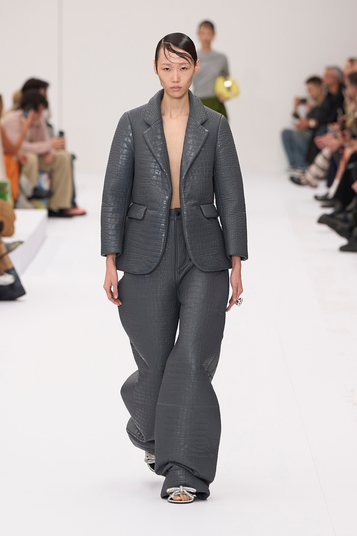 Model walks the runway in a textured charcoal grey crocodile-embossed suit with an oversized blazer and wide-leg pants; minimalist monochrome look.