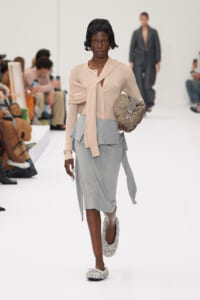 Runway model in a beige knit top with a long scarf, gray layered skirt, jeweled flats, and a quilted beige bag on her arm.