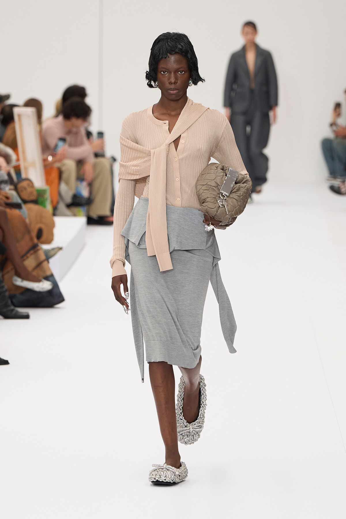 Runway model in a beige knit top with a long scarf, gray layered skirt, jeweled flats, and a quilted beige bag on her arm.