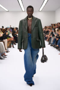 Runway model in a dark green crocodile-textured blazer over a bare chest, paired with wide-leg jeans and a chunky gold necklace, carrying a small patterned bag.
