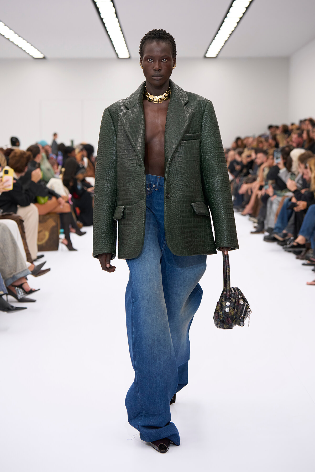 Runway model in a dark green crocodile-textured blazer over a bare chest, paired with wide-leg jeans and a chunky gold necklace, carrying a small patterned bag.