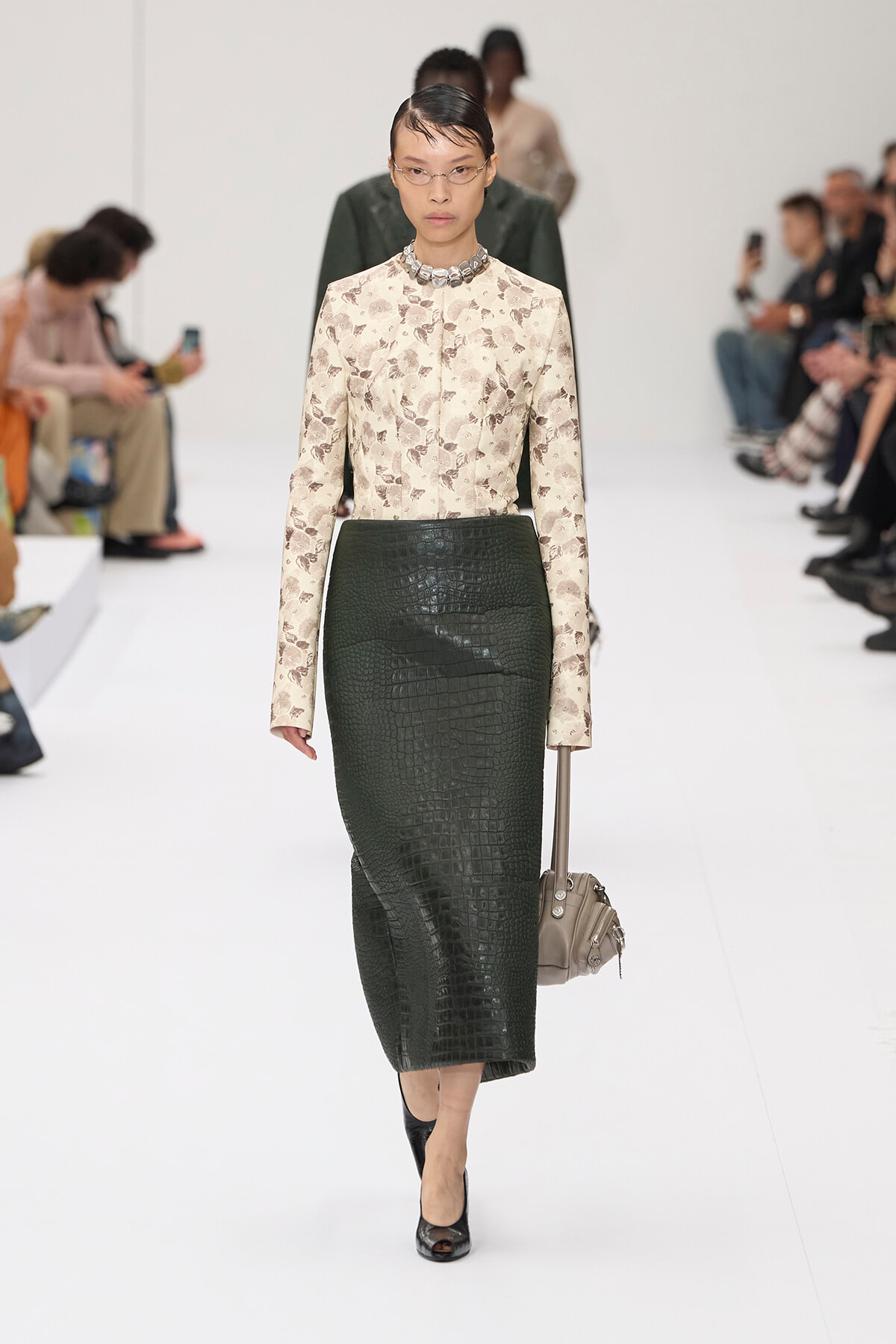 Model walking the runway in a cream floral top and dark green crocodile-textured pencil skirt, with a chunky silver necklace and taupe handbag.