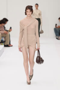 Model walking a beige knit dress with tied scarf on a white runway, holding a brown handbag and wearing peep-toe shoes.