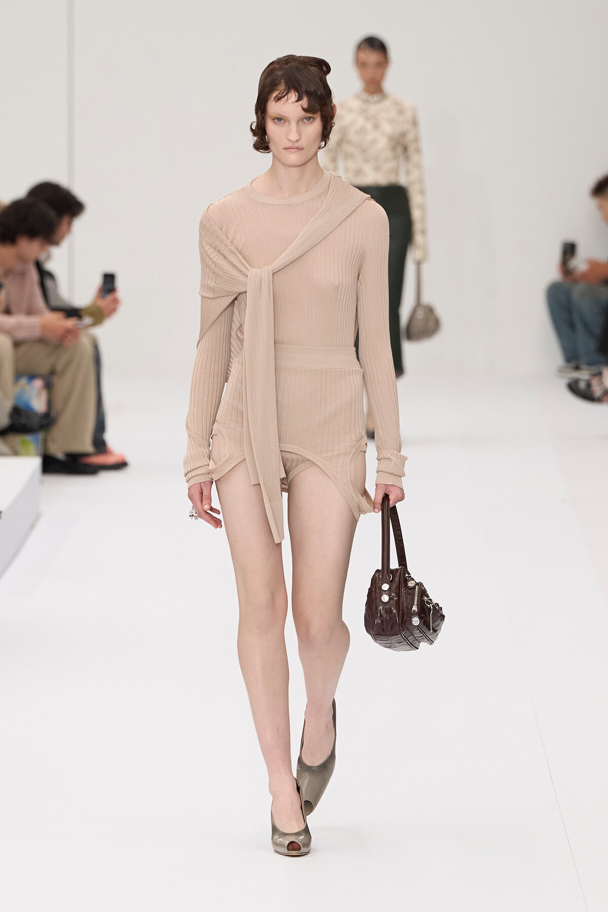 Model walking a beige knit dress with tied scarf on a white runway, holding a brown handbag and wearing peep-toe shoes.