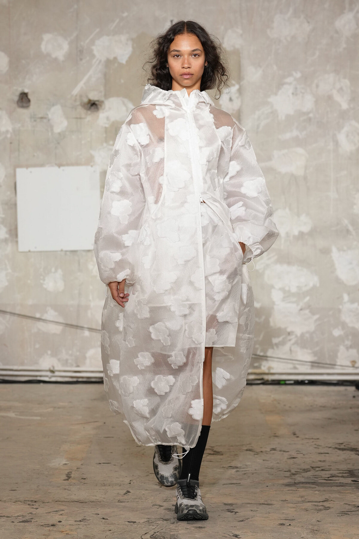 Female model walks a runway in a sheer white coat with cloud appliqués, oversized sleeves and a high collar, paired with black socks and chunky sneakers.