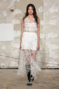 Model walking in a sheer white lace dress with floral appliqués, layered over a white slip, against an unfinished concrete backdrop.
