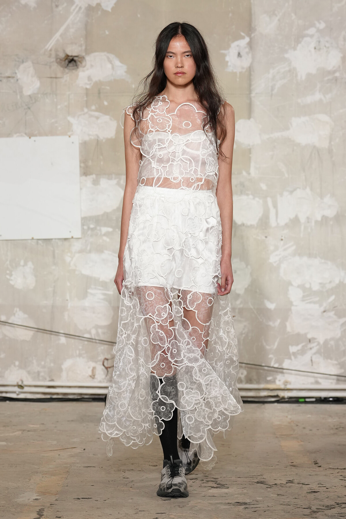 Model walking in a sheer white lace dress with floral appliqués, layered over a white slip, against an unfinished concrete backdrop.