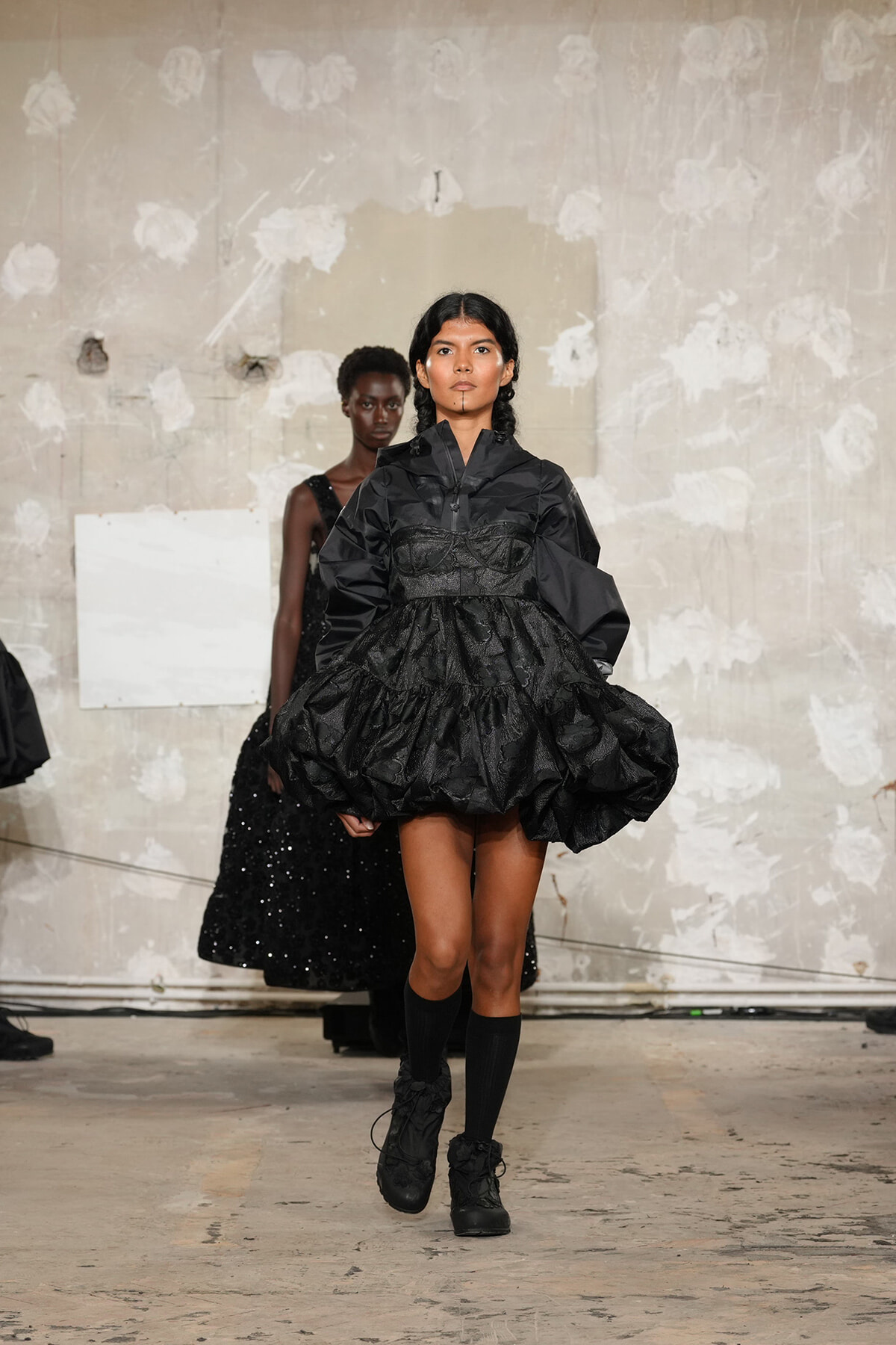Model walking a runway in a black, voluminous, textured dress with a structured bodice and high collar, paired with black boots.