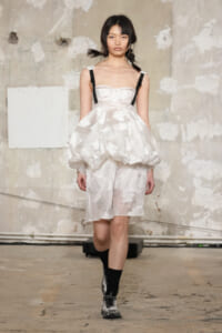 Model walks the runway in a white textured dress with a voluminous bubble skirt, black shoulder straps, and gray sneakers against an industrial backdrop.