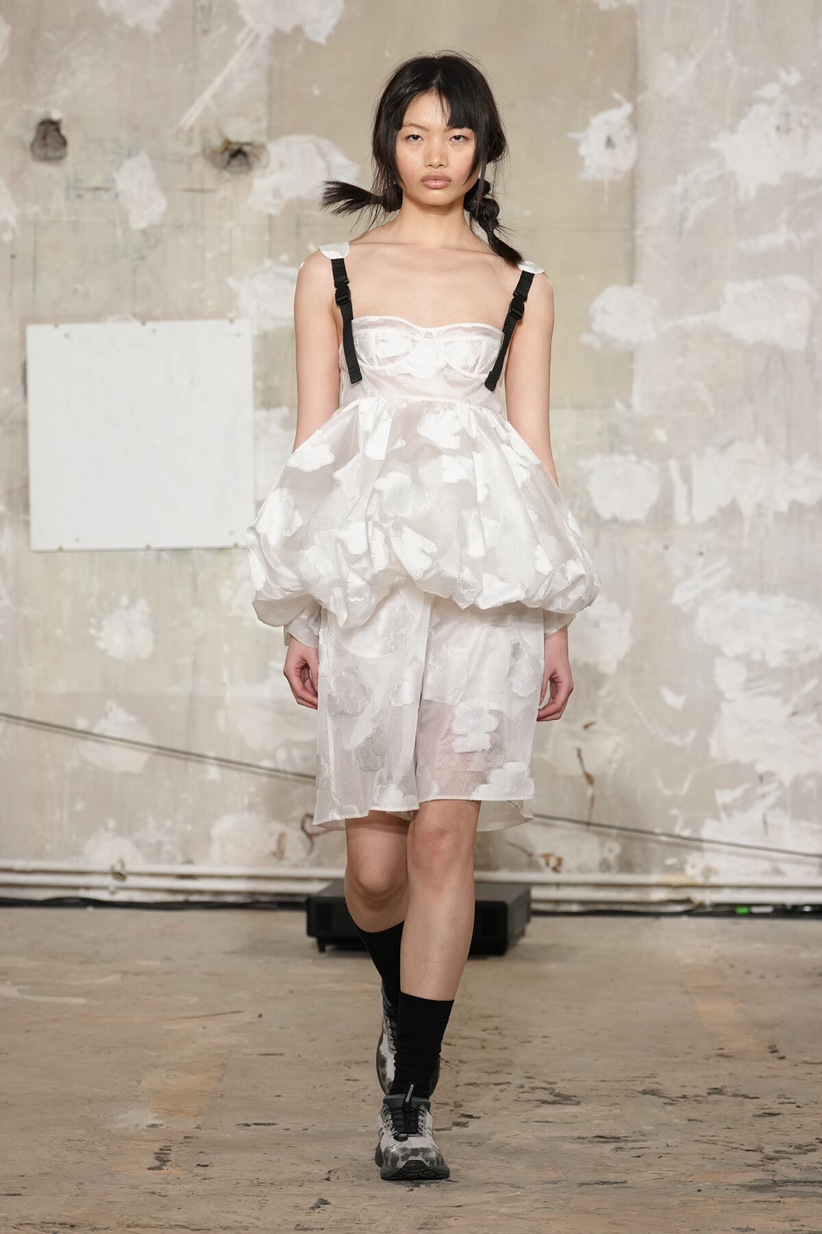 Model walks the runway in a white textured dress with a voluminous bubble skirt, black shoulder straps, and gray sneakers against an industrial backdrop.
