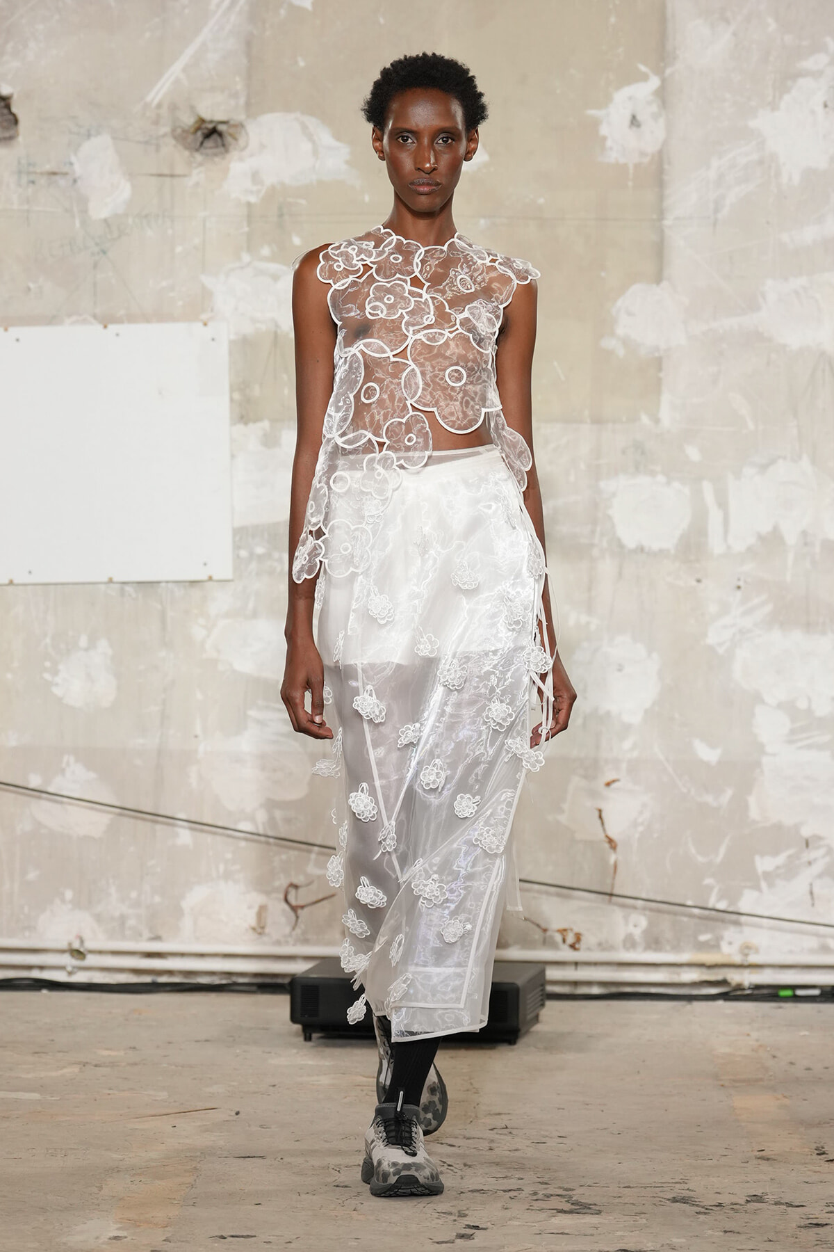 Model wearing a sheer, floral lace crop top with a matching white lace skirt in an industrial backstage setting.