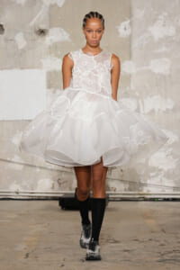 Model walking in a white organza dress with large floral appliqués on a concrete runway backdrop.