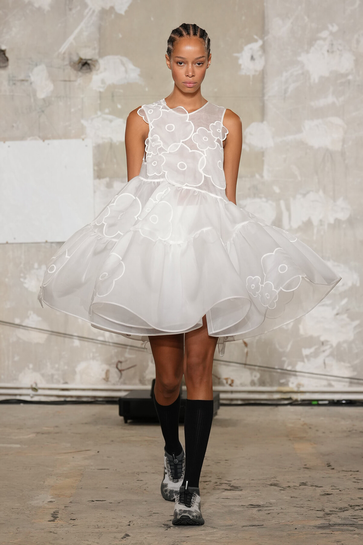 Model walking in a white organza dress with large floral appliqués on a concrete runway backdrop.