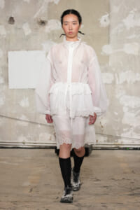 Model walks a runway in a sheer white zip-up jacket over a white dress with a floral lace hem, paired with black knee-high socks and camo sneakers.