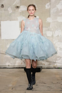 Model wearing a pale blue sheer dress with embroidered floral appliqués, walking on a fashion runway against an industrial backdrop.