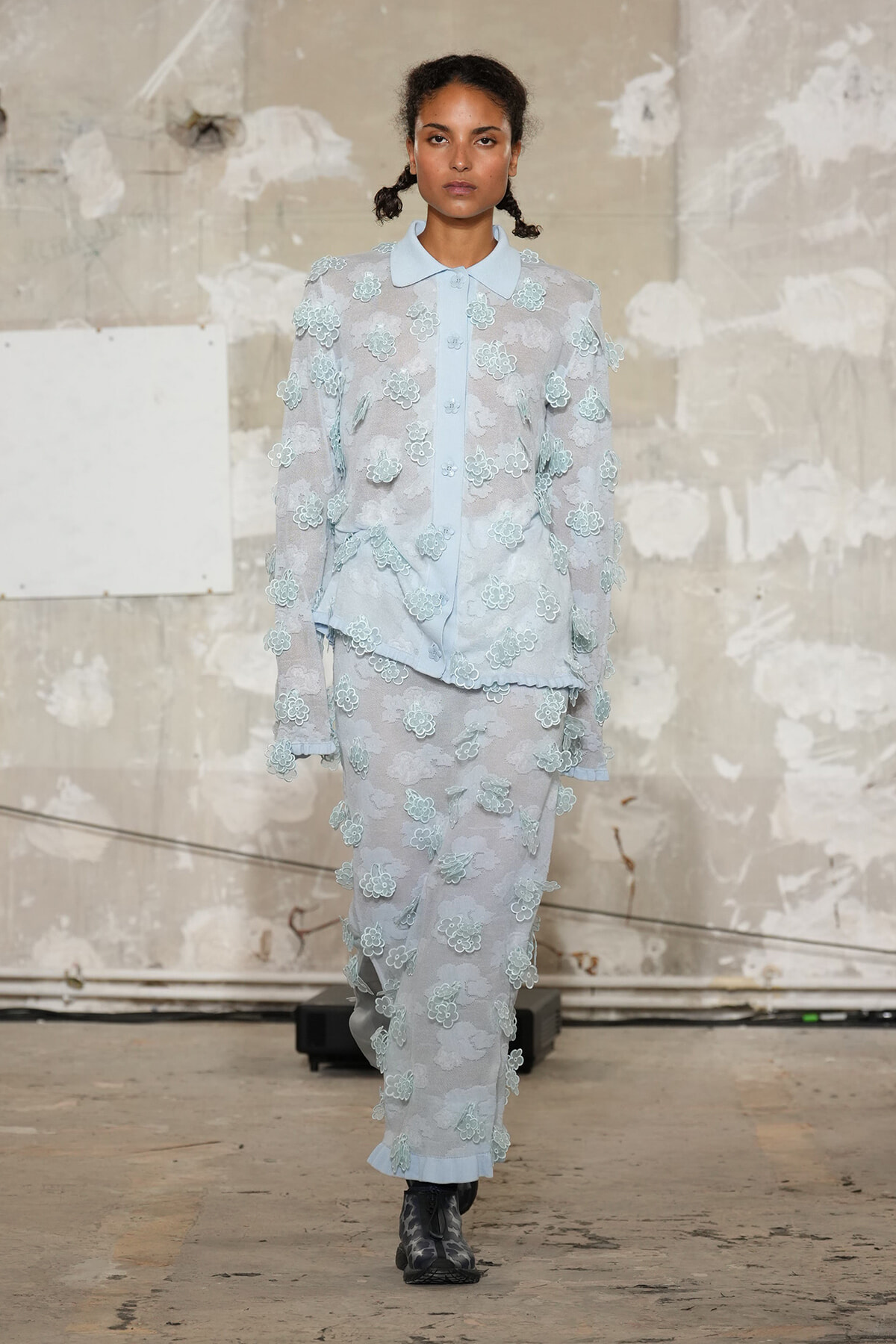 Model walking a fashion runway in a pale blue, sheer lace suit with 3D floral appliqués against a distressed studio backdrop.