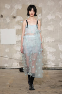 Runway model in a pale blue sheer dress with floral appliqués and ruffles, paired with black strap accents and boots.