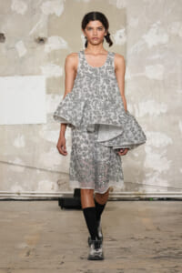 Model walking a runway in a silver sequin floral dress with a dramatic layered cape-like overlay in an industrial studio setting.