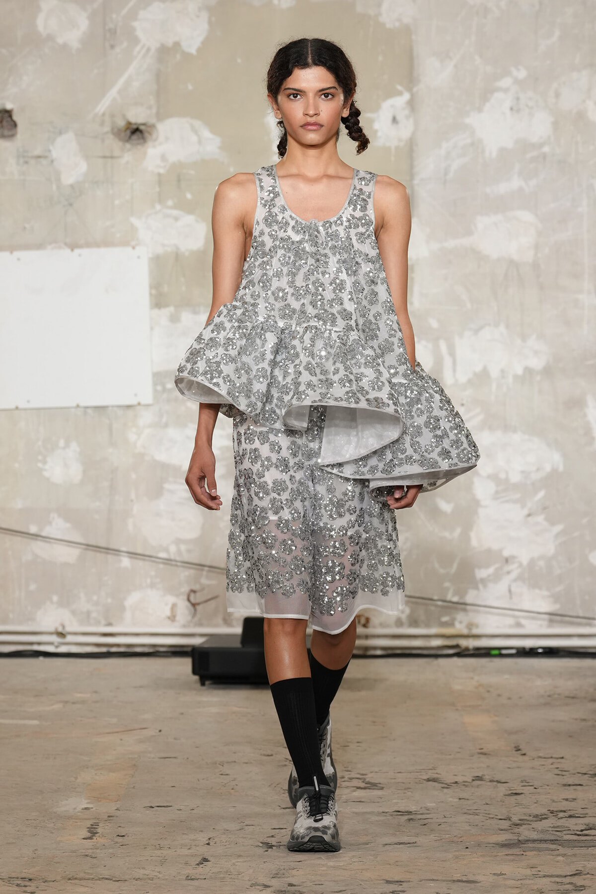 Model walking a runway in a silver sequin floral dress with a dramatic layered cape-like overlay in an industrial studio setting.