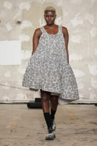 Model walking the runway in a silver sequin A-line dress; industrial backdrop in view.