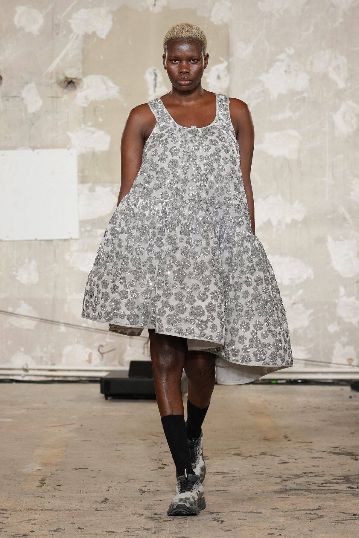 Model walking the runway in a silver sequin A-line dress; industrial backdrop in view.