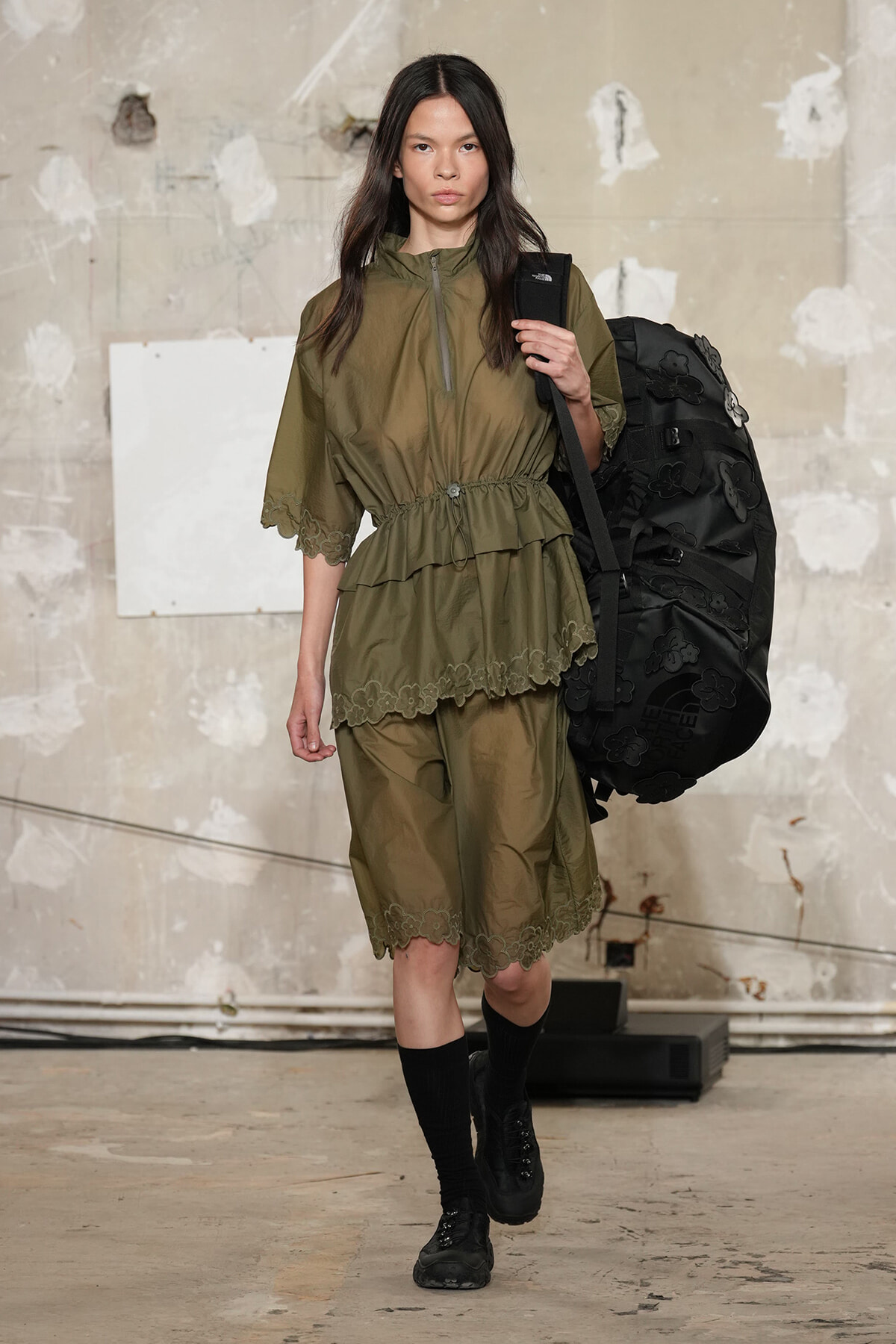 Model wearing an olive-green utility tunic with a cinched waist and matching shorts with lace trim, carrying a large black backpack in an industrial setting.