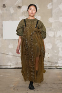 Model wearing olive green puff-sleeve top underneath a brown sheer dress with floral embroidery and ruffles, walking a runway.