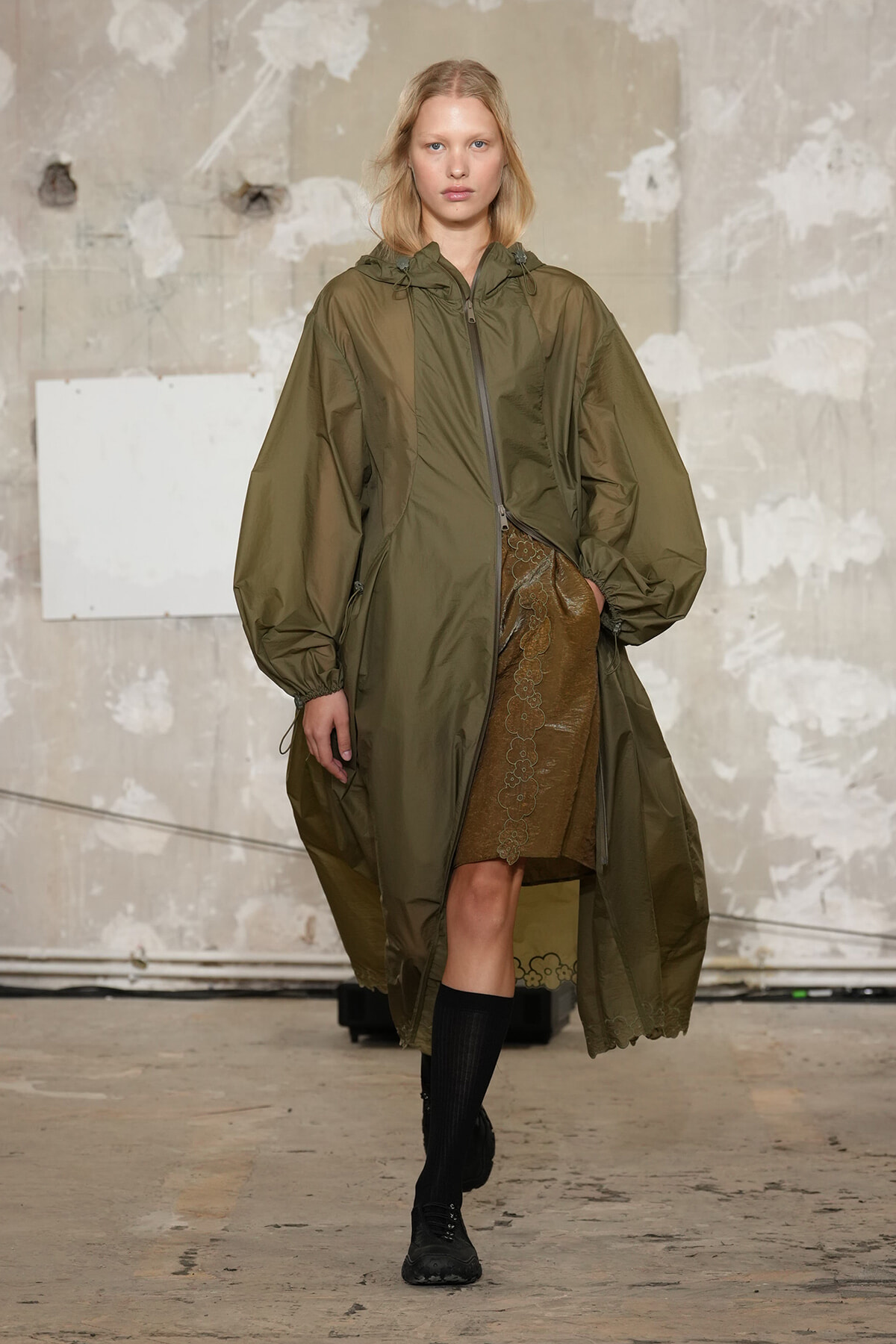 Model wears an oversized olive parka with a hood and front zipper, over a brown lace‑trimmed skirt with black knee‑high socks on a runway backdrop.