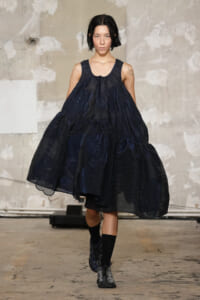 Model walking a dark navy, voluminous dress on a fashion runway, striking pose and short black hair.