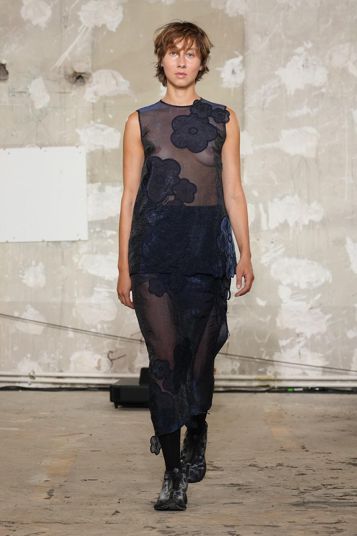 Female model walks a fashion runway wearing a sheer navy blue dress with large floral appliqués and dark underlayers.
