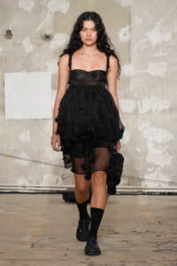 Female model walking a fashion runway in a black layered dress with floral appliqués, sheer overskirt, and chunky-strap bodice, paired with black socks and shoes in an industrial setting.