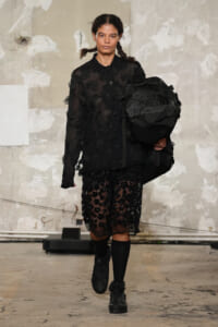 Female model on a runway wearing a sheer black lace dress with floral appliqués, carrying a large black The North Face duffel bag over her shoulder.