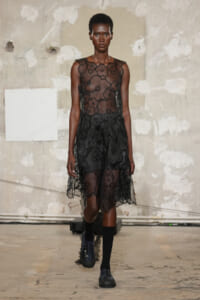 Model walks the runway in a sheer black floral lace dress with a translucent bodice and layered skirt.