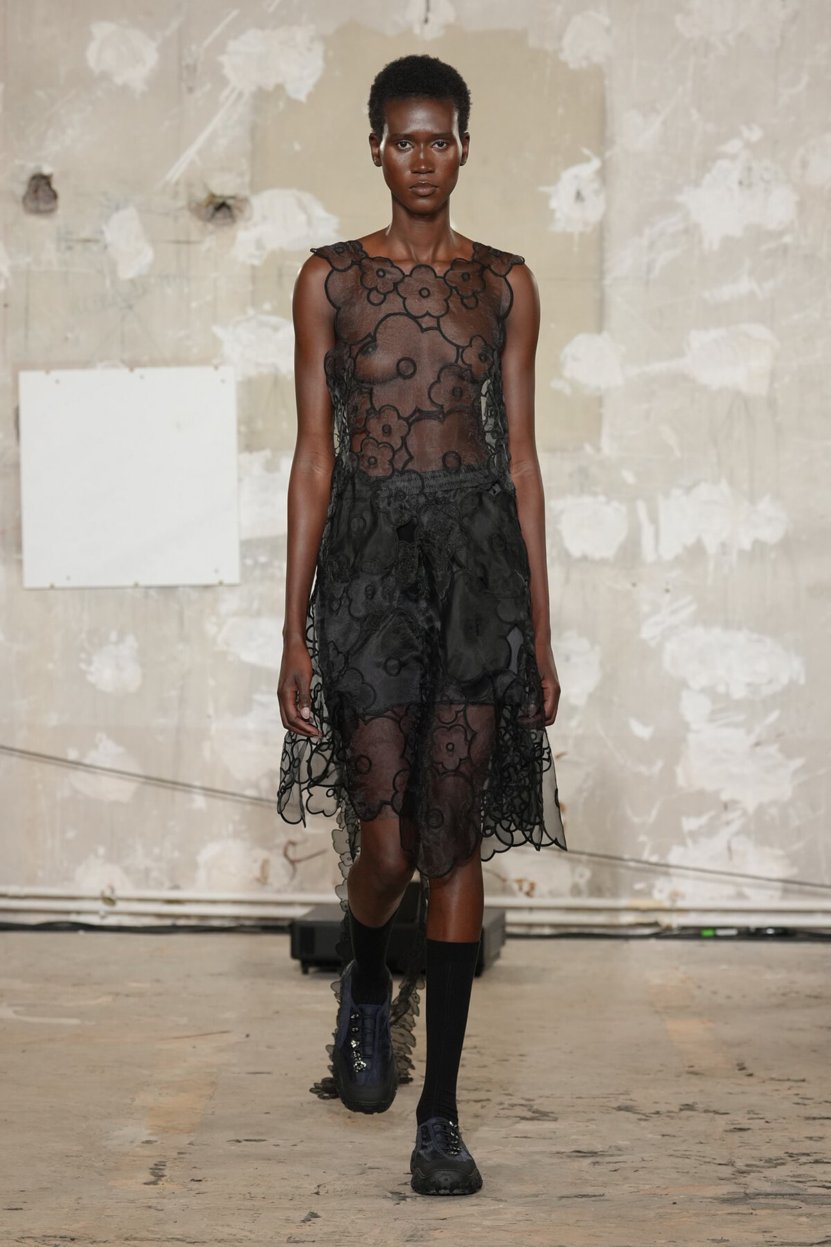 Model walks the runway in a sheer black floral lace dress with a translucent bodice and layered skirt.