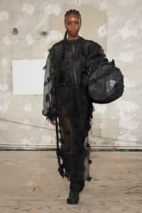 Model walking a runway in a sheer black dress with floral appliqués, carrying a large black North Face duffel bag.