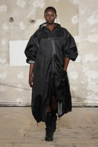 Female model walking a runway in a long black parka with subtle floral embroidery and hood, industrial backdrop behind.