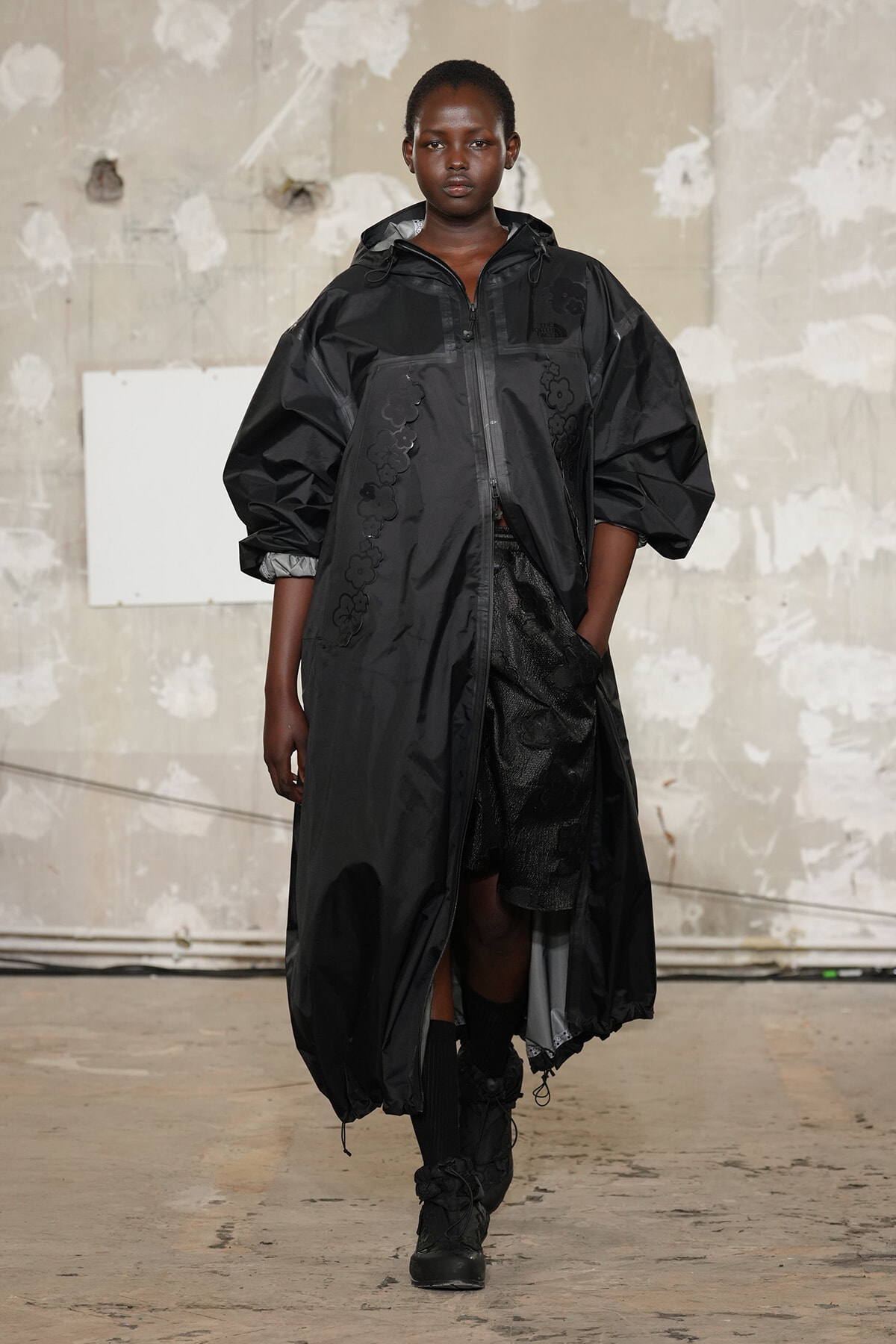 Female model walking a runway in a long black parka with subtle floral embroidery and hood, industrial backdrop behind.