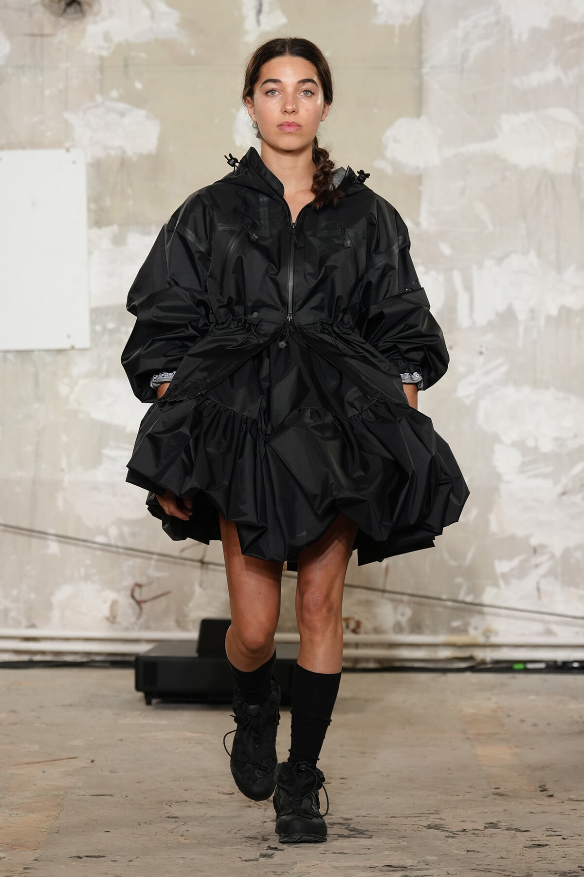Model walking a runway in an oversized black jacket with a gathered waist and large ruffled hem, black knee-high socks and boots.
