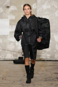 Model walks a runway in a black oversized windbreaker with a drawstring waist, carrying a large black floral-patterned bag over her shoulder.