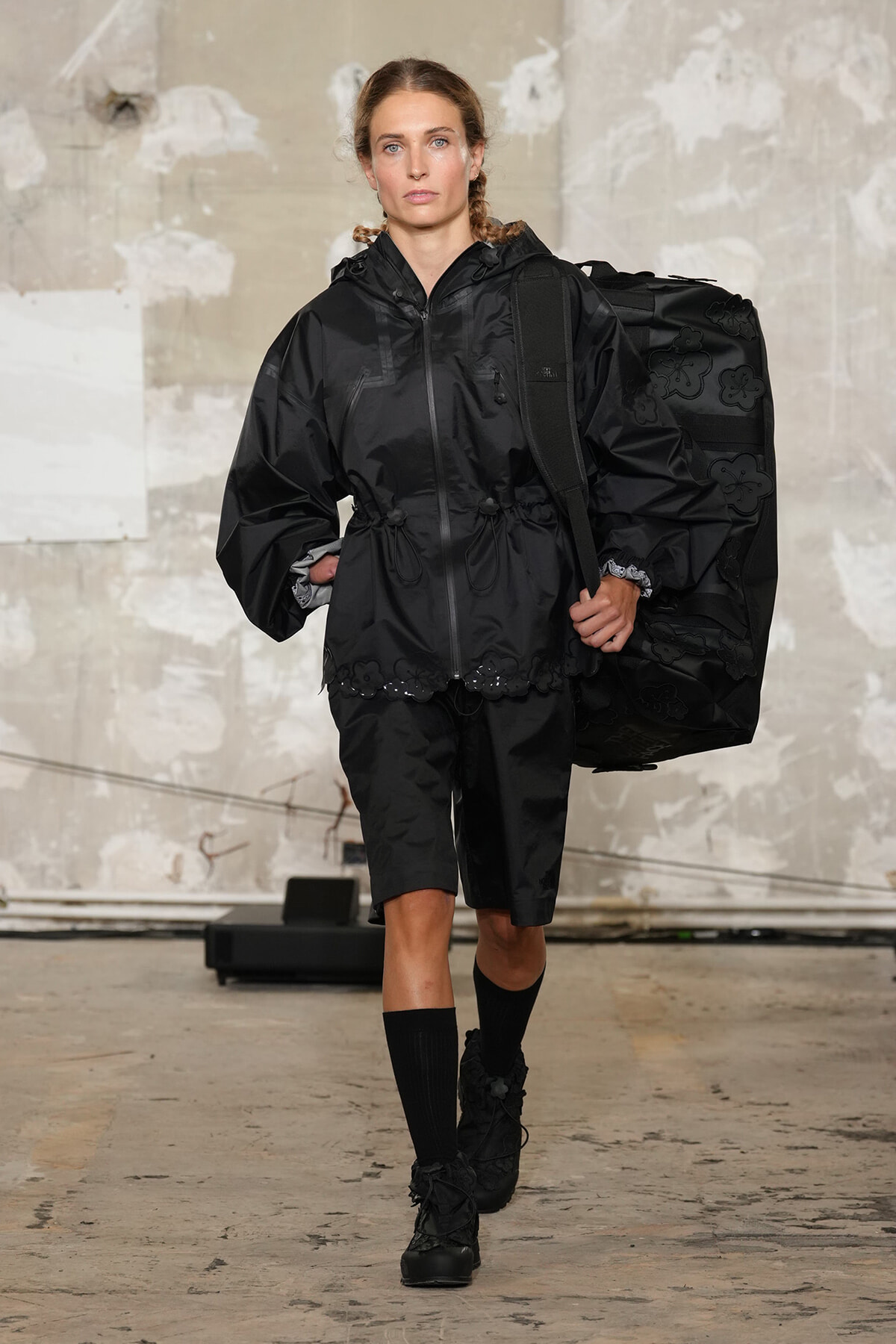 Model walks a runway in a black oversized windbreaker with a drawstring waist, carrying a large black floral-patterned bag over her shoulder.