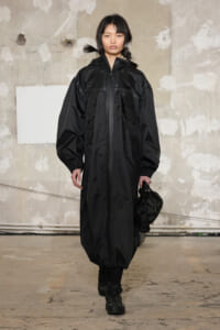 Model walking in a long black hooded windbreaker with elastic cuffs, holding a black bag in one hand in a bare industrial space.
