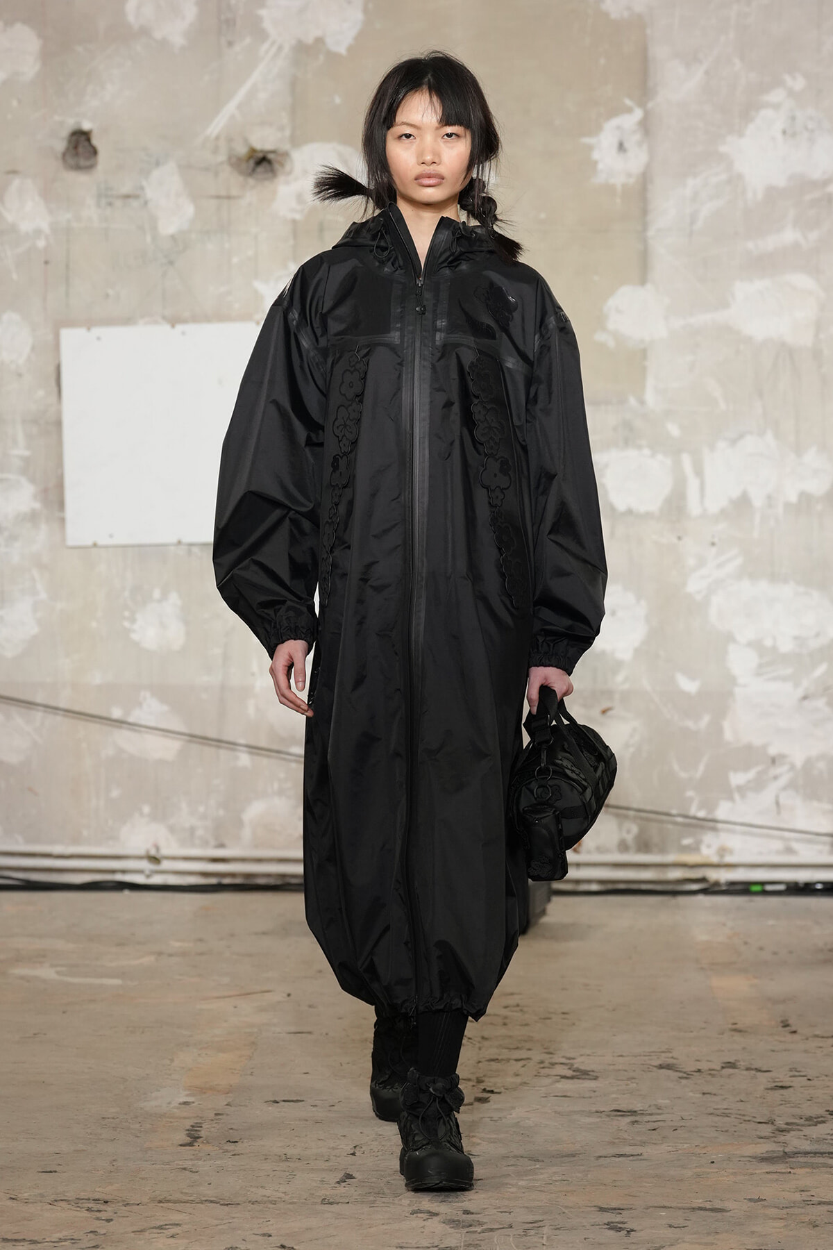 Model walking in a long black hooded windbreaker with elastic cuffs, holding a black bag in one hand in a bare industrial space.