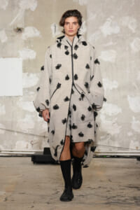 Runway model in a loose gray coat with black floral appliqués and matching panels, paired with black boots.