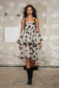Model walking runway in a light grey dress with black floral appliqués and thick black straps, pairing knee-high black socks and boots for a fashion show look.