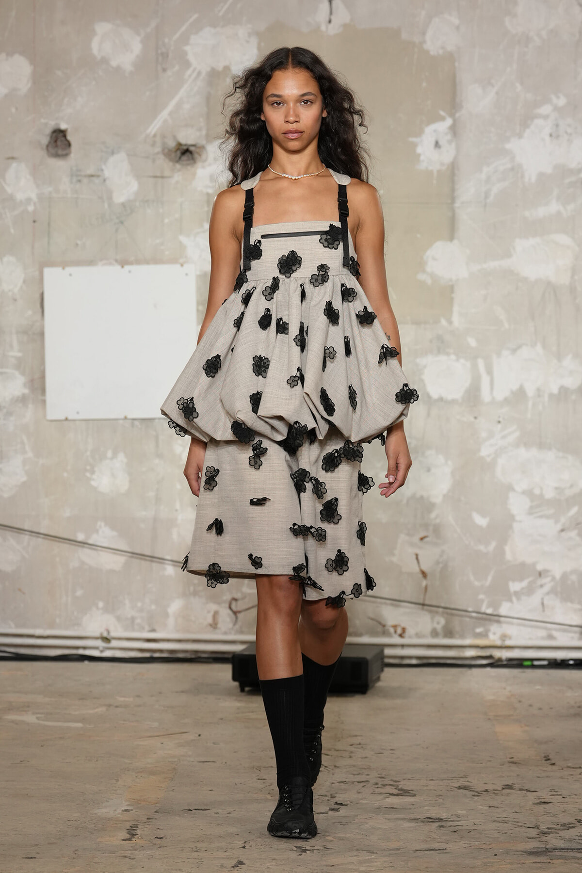 Model walking runway in a light grey dress with black floral appliqués and thick black straps, pairing knee-high black socks and boots for a fashion show look.