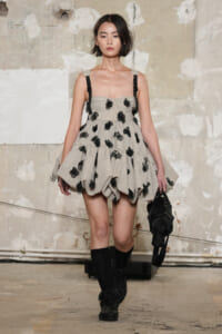 Model on a runway wearing a beige voluminous dress with black floral appliqués, black straps, and knee-high socks, carrying a black bag.