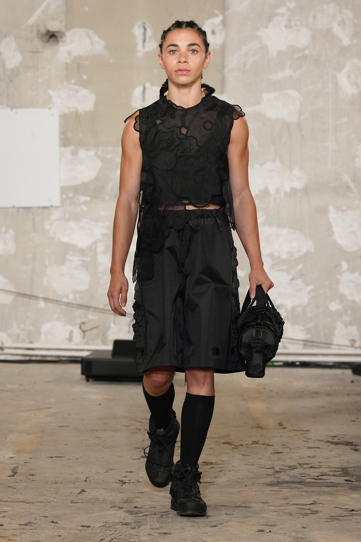 Female runway model in an all-black ensemble: floral lace sleeveless crop top with matching shorts, knee-high socks, and black boots, carrying a black handbag.