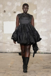 Model walking the runway in a black, tiered puff-sleeve dress with a lace bodice and knee-high socks against an industrial backdrop.