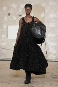 Model walks the runway in a floor-length black sequined gown and chunky boots, carrying a large black backpack with flower patches.