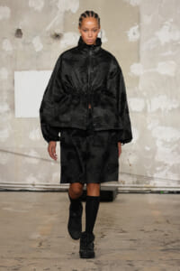 Model walking a runway in a black, textured floral set: oversized zip-front jacket cinched at the waist and matching knee-length shorts with black socks and boots.